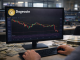 A trader’s hands operate a keyboard while monitoring a Dogecoin candlestick chart on a desktop monitor in a financial office setting.